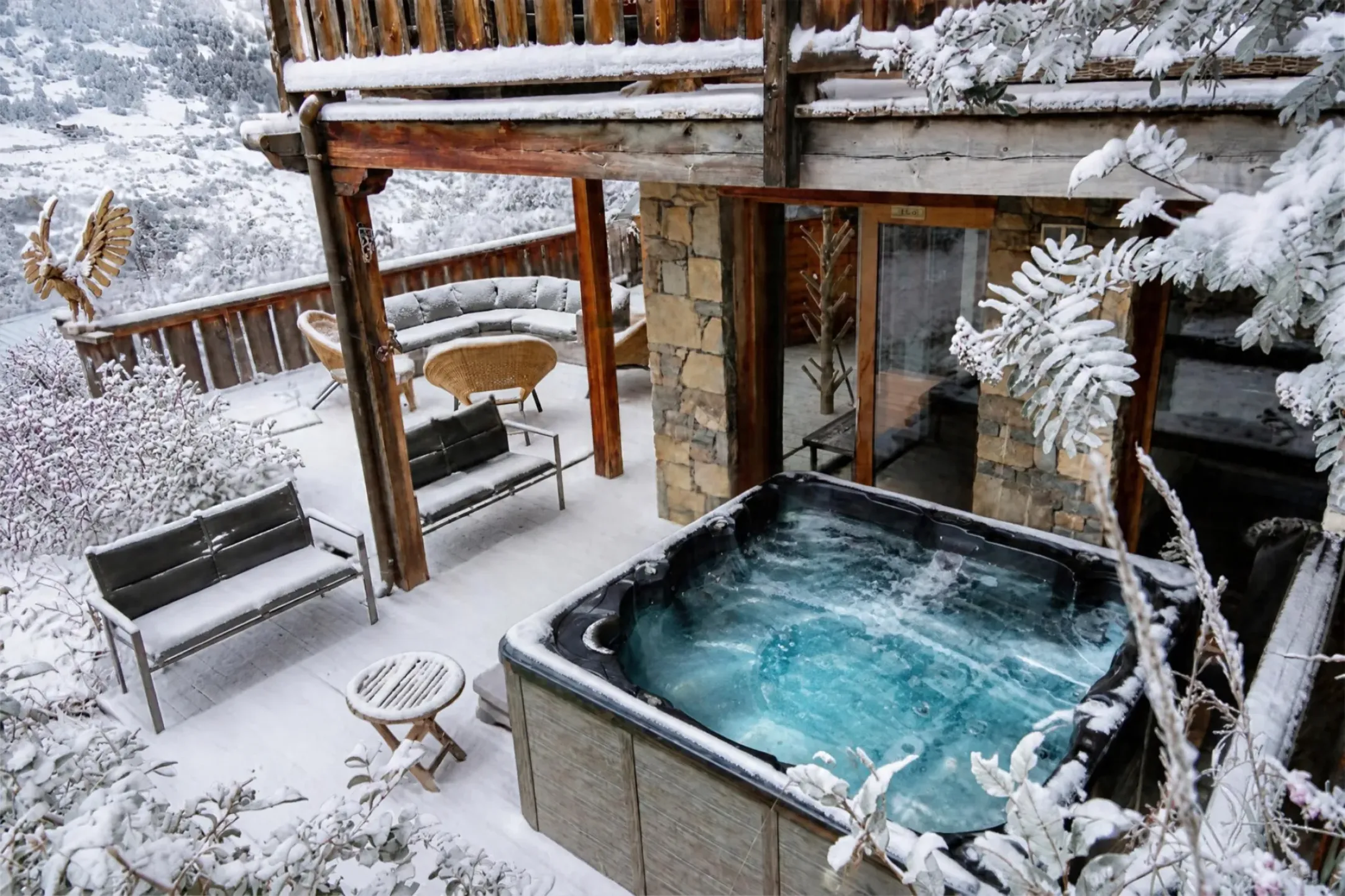 Terrasse du Chalet D à Vars recouverte de neige, jacuzzi extérieur à l'eau turquoise, bancs, salon de jardin et fauteuils en rotin enneigés, sculpture d'aigle en bois, arbustes givrés et vallée blanche en arrière-plan